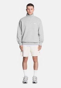 Męski model stoi na tle białym, ubrany w szaro-suwakową bluzę i białe szorty, prezentując sportowe obuwie i tatuaże.