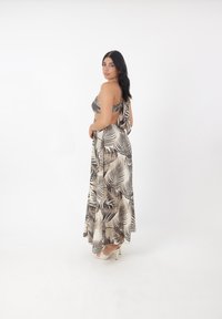 Schwarz-beige Maxi-Kleid mit Palmenblatt-Druck, tailliertem Oberteil, offener Rückseite und fließendem Rock. Kombiniert mit hellen, hohen Sandalen.