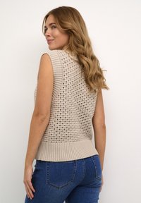 Pull en tricot sans manches beige avec un design texturé et un tricot ajouré, doté d'un ourlet côtelé. Associé à un jean bleu.