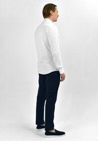 Witte lange mouwen overhemd met een gebogen zoom, gecombineerd met zwarte slim-fit broek en zwarte instap schoenen met witte zolen. Neutrale achtergrond.