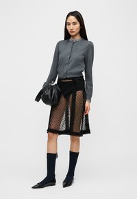 Cardigan gris à boutons, jupe noire transparente à pois avec bords volantés, sac noir, chaussettes foncées et chaussures pointues noires.