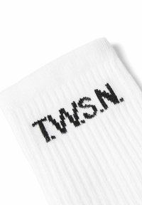 Hvid ribbet stof, nærbillede der viser sorte broderede bogstaver "T.W.S.N." i hjørnet.