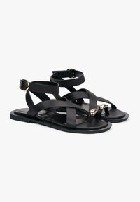 Zwarte platte sandalen met gekruiste bandjes van een glad materiaal. Bevat gouden accenten bij de teen en de enkelband voor sluiting.