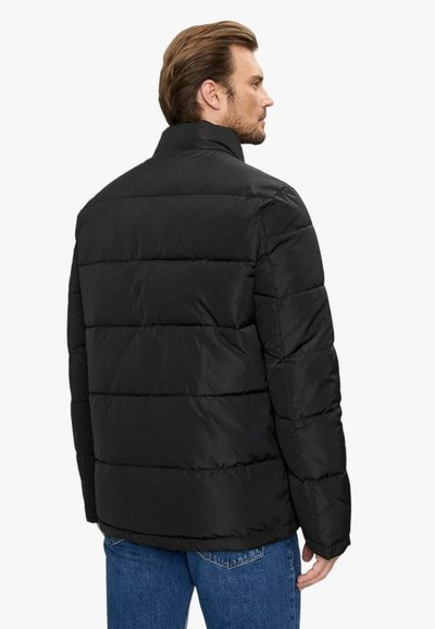 Giubbotto trapuntato nero con cuciture orizzontali, colletto alto e una texture liscia. Presenta una vestibilità rilassata ed è indossato sopra dei jeans blu.