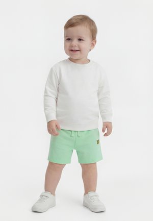 Tout-petit aux cheveux châtain clair portant un t-shirt blanc à manches longues, un short vert menthe avec un logo jaune, et des baskets blanches, debout et souriant.