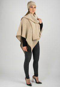 Poncho lavorato a maglia beige con un motivo a zigzag, abbinato a un cappello coordinato. Texture liscia, design a strati, indossato sopra una maglietta nera e jeans.