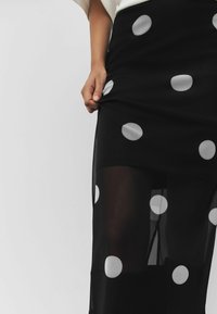 Zwarte doorschijnende rok met grote witte stippen, met een rechte vorm en lichte textuur. Het model houdt de stof bij de taille vast.