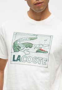 Άνδρας που φοράει λευκό μπλουζάκι με πράσινο και ροζ κεντημένο κροκόδειλο και το κείμενο "LACOSTE" σε φόντο πλέγματος.