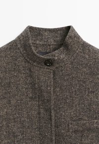 Brown herringbone-patterned fabric with a high collar and a single black button closure. Features a subtle texture and tailored design.