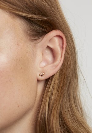 Goldene blumenförmige Ohrringe mit drei Edelsteinen: einem hellen Pfirsichton, einem blauen Zentrum und einem klaren Akzent. Nahaufnahme an einem Ohr.