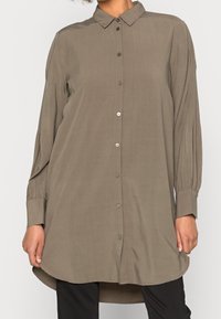 Chemise à manches longues, couleur olive avec un devant boutonné, un col classique et un ourlet arrondi. Présente une légère texture et des manches à poignets.