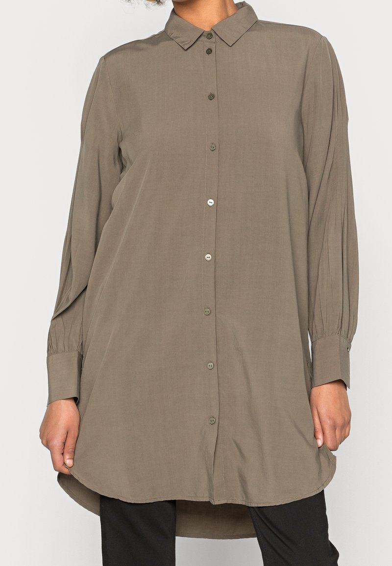Chemise à manches longues, couleur olive avec un devant boutonné, un col classique et un ourlet arrondi. Présente une légère texture et des manches à poignets.