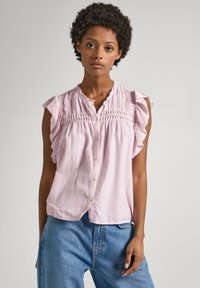 Blouse rose clair à manches courtes avec des accents de volants, corsage smocké, fermeture à boutons et coupe décontractée, associée à un jean bleu décontracté.