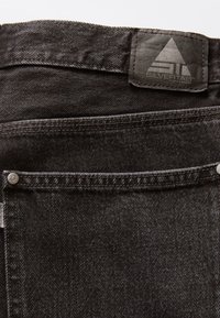 Svarta denimjeans har en strukturerad yta, synlig sömnad och en läderlogotagsruta vid midjan. Silverfärgade detaljer finns.