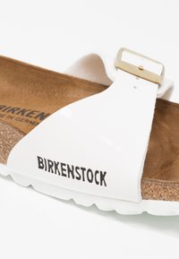 Witte Birkenstock sandaal met een gladde bovenkant, verstelbare metalen gespsluiting, bruine kurken voetbed en geribbelde witte zool met logodetail.
