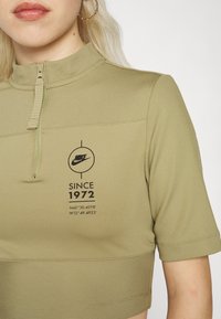 Olivově zelený zkrácený zip-up top vyrobený z žebrované tkaniny, s vysokým límcem a kruhovým logem s textem: "SINCE 1972" a souřadnicemi.