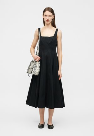 Femme portant une robe midi noire sans manches, un sac à épaule à motif peau de serpent, et des ballerines noires, debout sur un fond blanc.