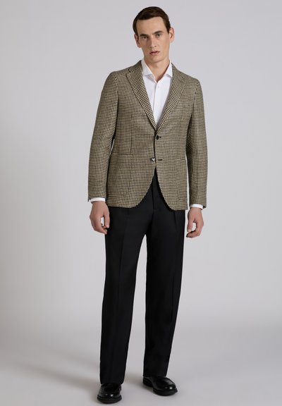 Ein Mann steht vor einem schlichten grauen Hintergrund und trägt ein braun-weißes Hahnentritt-Sakko, ein weißes Hemd, schwarze Hose und schwarze Schuhe.