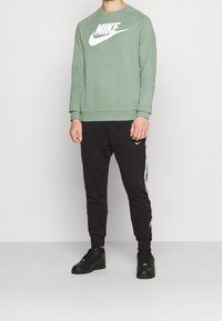 Zelená mikina Nike s bílým logem, v kombinaci s černými tepláky s bílými bočními pruhy a černými teniskami. Neformální střih.