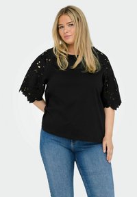 Black cotton t-shirt with lace sleeves featuring floral cut-out patterns. Round neckline and relaxed fit. Paired with blue jeans.