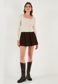 Grietas juostelėmis megztinis su apvaliu iškirpimu ir mygtukais, derinamas su rudais klostuotais mini sijonais ir iki kelių siekiančiais rudais batais su auksiniais akcentais.