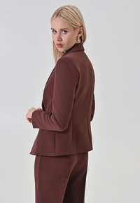 Brauner maßgeschneiderter Blazer mit strukturierter Passform, langen Ärmeln und Reverskragen, kombiniert mit passender Hose, glatte Textur, minimale Details.