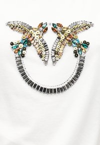 Δύο στολισμένα πουλιά που κοιτάζονται μεταξύ τους πάνω από μια καμπυλωτή σειρά από ορθογώνιες στρας σε λευκό ύφασμα.