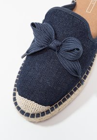 Espadrilles en denim bleu marine avec un détail de nœud tressé, semelle en jute naturel et coutures contrastées le long du bord. Texture lisse et forme arrondie.