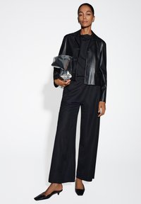 Zwarte leren cropped jas met knopen, gecombineerd met een zwarte top en wijde broek. Houdt een metalen clutch vast, afgemaakt met zwarte puntige flats.