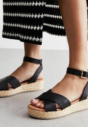 Czarne skórzane sandały na koturnie z krzyżującymi się paskami i zapięciem na kostkę, z naturalną plecioną juty podeszwą i gładką brązową wkładką.