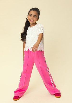 Girl smiling, standing with legs apart, wearing a white eyelet blouse and bright pink pants with heart-patterned stripes and red shoes.