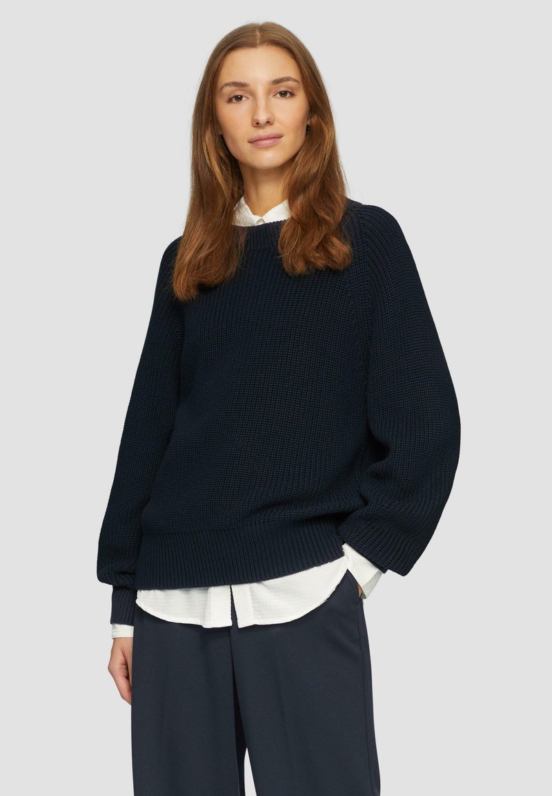 Maglione lavorato a maglia blu navy con orlo a coste e maniche ampie, indossato sopra una camicia bianca con colletto, abbinato a pantaloni scuri.