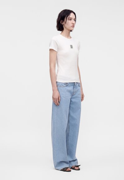Jeune femme debout portant un t-shirt blanc à manches courtes avec un logo et un jean bleu clair à jambes larges avec des sandales noires à bout ouvert.