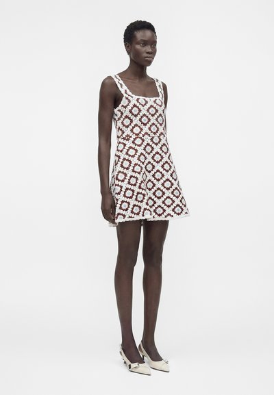 Μαύρη γυναίκα στέκεται φορώντας ένα αμάνικο, πλεκτό φόρεμα μήκους μέχρι το γόνατο με καφέ και λευκό τετράγωνο λουλουδάτο σχέδιο και λευκές μυτερές γόβες.