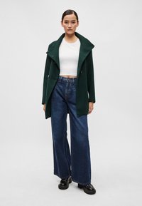 Casaco de lã verde com uma lapela larga e botões duplamente abotoados, usado sobre um top cropped branco e combinado com jeans azuis de perna larga de cintura alta.