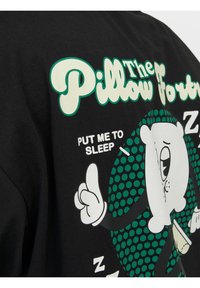 Černé tričko s krátkým rukávem, které zobrazuje design kreslené medvěda s textem "Pillow Fort" a "PUT ME TO SLEEP" v kontrastních barvách.