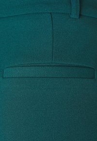 Teal fabric features a smooth texture with a flat pocket detail at the back. The design is tailored with a clean finish and subtle stitching.