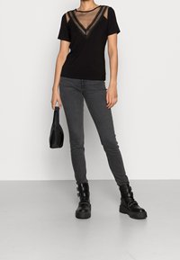 Kobieta w czarnej siateczkowej bluzce z dekoltem w serek, szarych obcisłych dżinsach, czarnych botkach z klamrą, trzymająca małą czarną torebkę, stojąca przy gładkiej białej ścianie.