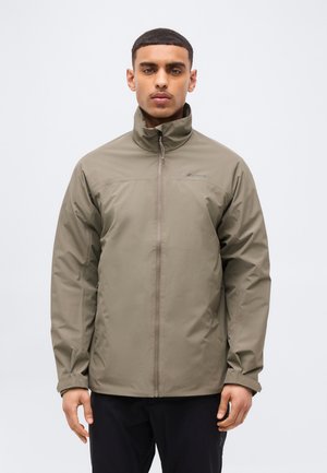 Lühikeste juustega mees, kes kannab oliivrohelise lukuga jopet ja musti pükse, seisab ühetaolise heleda tausta ees.