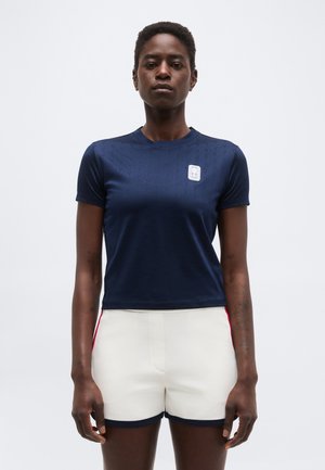 Ženska stoji obrnjen naprej, oblečena v oprijeto mornarsko modro majico z logotipom na prsih in bele kratke hlače z rdečimi stranskimi črtami.