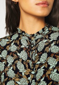 Blusa floreale con sfondo nero, fiori verde chiaro e foglie marroni. Collo arricciato e dettaglio con bottone dorato.