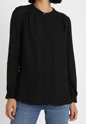 Femme portant un chemisier noir à manches longues avec un col rond et une fermeture par un seul bouton, associé à un jean bleu.