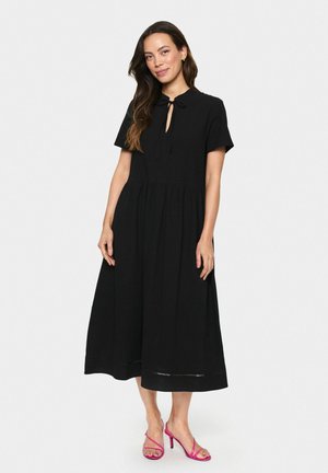 Zwart midi-jurk met korte mouwen, sleutelgat halslijn en een geplooide taille. Gladde stof, losse pasvorm, gecombineerd met roze haksandalen.