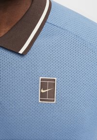 Blå mesh polo skjorte med brun krage og hvite striper, med et kvadratisk merke med en gull Nike-logo.