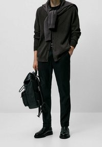 Cardigan marron foncé sur une chemise noire, pantalon noir fuselé, sac à dos en cuir et bottines noires. Textures lisses et accessoires minimalistes.