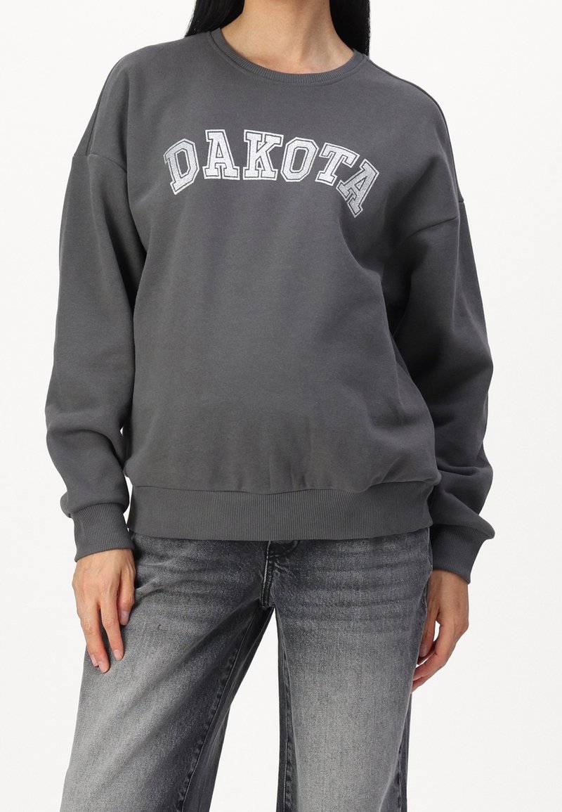 Personne portant un sweat-shirt gris foncé avec « DAKOTA » en lettres blanches et un jean noir délavé, debout devant un fond uni.