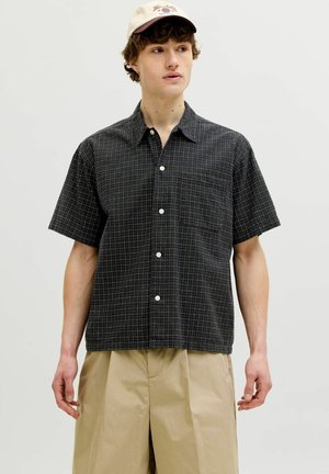 Ung mann som har på seg en svart rutete kortermet skjorte med knapper, beige bukser og en beige baseballcaps, står foran en ensfarget bakgrunn.