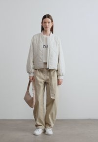 Καπιτονέ ανοιχτό γκρι μπουφάν τύπου bomber, λευκό πουλόβερ με κείμενο, φαρδιά μπεζ παντελόνια και λευκά αθλητικά παπούτσια. Κρατάει μία ταμπά τσάντα ώμου.