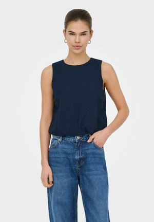 Kvinde med brunt hår sat op i en knold, iført guldringøreringe, en ærmeløs marineblå top og højtaljede blå jeans, poserer med den ene hånd i lommen.