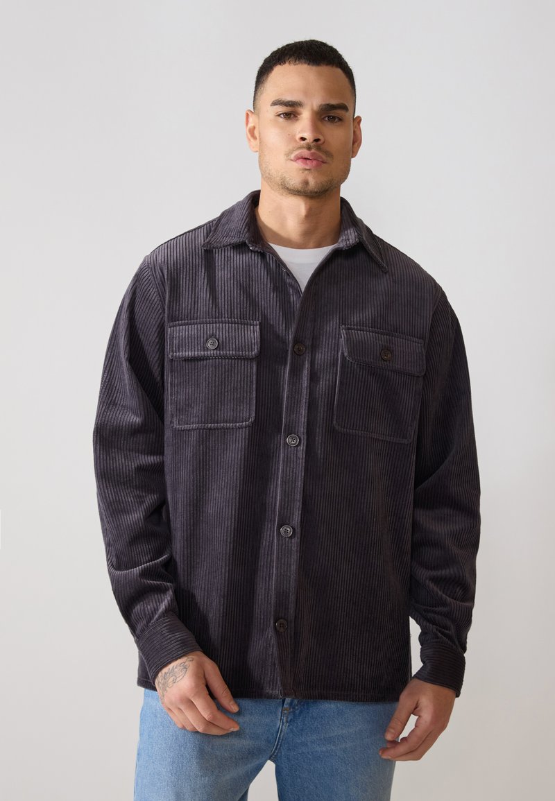 Chemise en velours côtelé gris avec deux poches poitrine, fermeture à boutons et col. Présente une surface texturée et une coupe décontractée.
